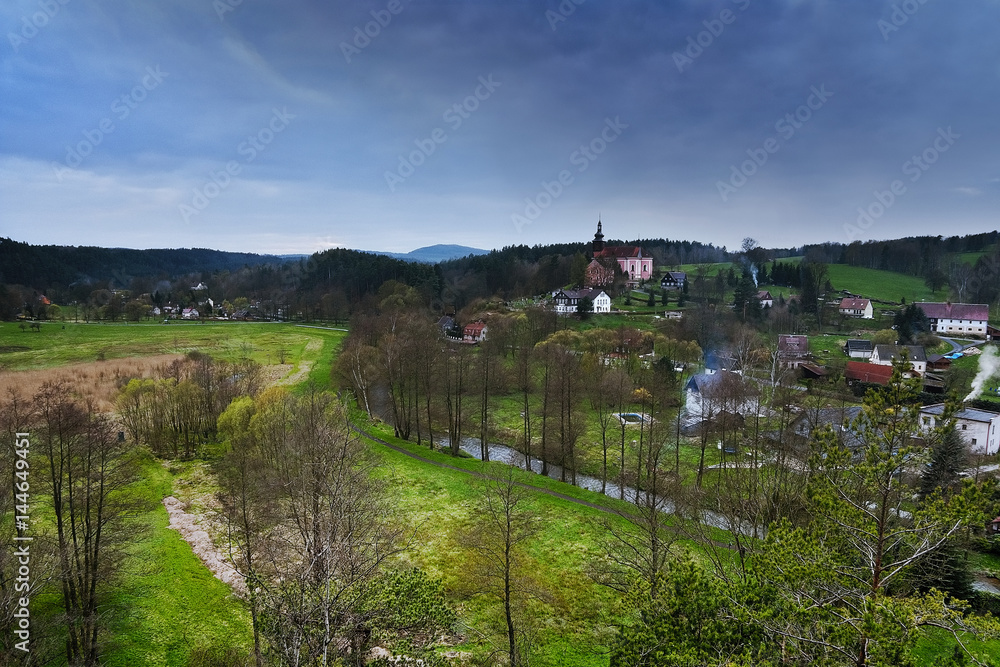 Srbska Kamenice, Czech republic - April 08, 2017: river Kamenice flowing through village with a church on hill in spring  natural reserve Arba in tourist area Labske piskovce at evening