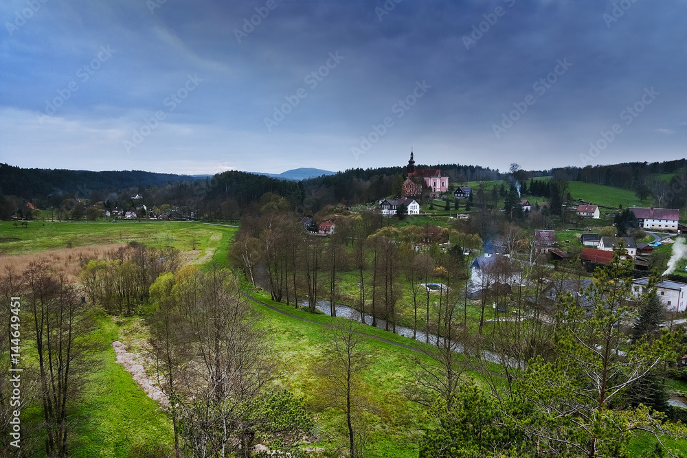 Srbska Kamenice, Czech republic - April 08, 2017: river Kamenice flowing through village with a church on hill in spring natural reserve Arba in tourist area Labske piskovce at evening Srbska Kamenice, Czech republic - April 08, 2017: river Kamenice flowing through village with a church on hill in spring natural reserve Arba in tourist area Labske piskovce at evening