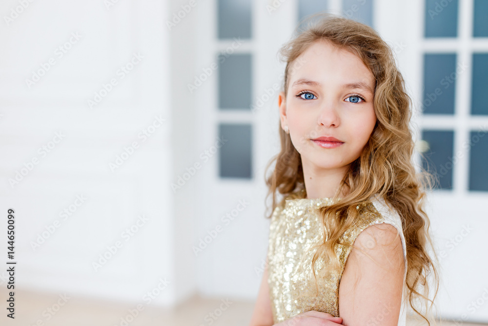 A portrait of a beautiful girl with long curly blond hair and blue eyes wearing a gold fashionable dress and high-heeled shoes