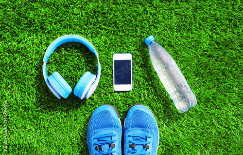 Blue a headphones and white smartphone with sports sneakers shoes, bottle of water on a green grass textured background, top view, flat lay photo