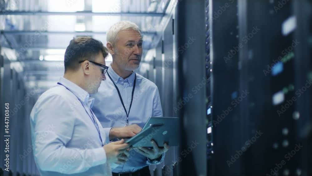 Two IT Technicians Standing in Data Center. They Use Laptop and Tablet Computer while Standing Beside Open Server Rack Cabinet. Shot on RED EPIC-W 8K Helium Cinema Camera. Two IT Technicians Standing in Data Center. They Use Laptop and Tablet Computer while Standing Beside Open Server Rack Cabinet. Shot on RED EPIC-W 8K Helium Cinema Camera.