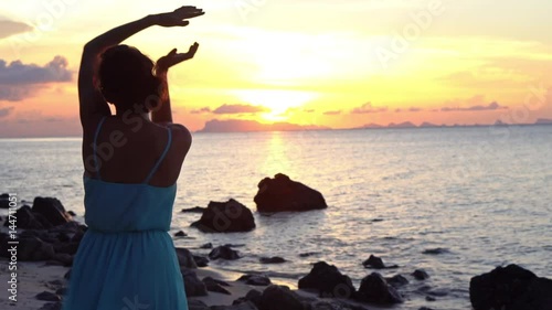 Happy young woman in a long dress dancing on the beach on a colourful sunset background
