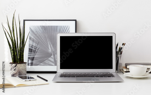 Creative designer desktop with blank copy screen laptop, bw photo in a frame, smartphone, stationery, green plant in a pot, cup of coffee on white wall background. Stylish hipster workspace, mock up