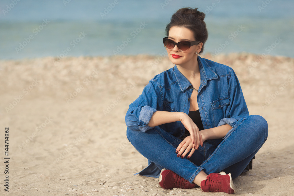 Denim outfit fashion details. Relaxing stylish woman enjoying the sun with red glitter manicure in navy jeans holding sunglasses and leather small cross body bag.
