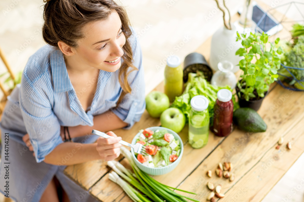 Plakat Młoda i szczęśliwa kobieta je zdrowego sałatkowego obsiadanie na stole z zielonymi świeżymi składnikami indoors