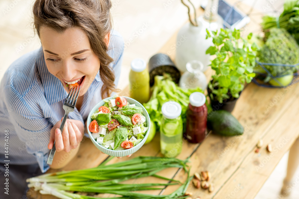 Plakat Young and happy woman eating healthy salad sitting on the table with green fresh ingredients indoors