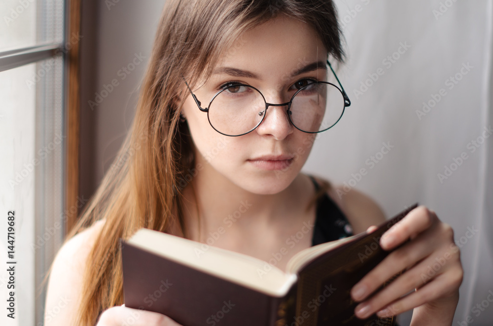 Beautiful girl sitting at the window, holding a book in his hands and looking at the camera Beautiful girl sitting at the window, holding a book in his hands and looking at the camera