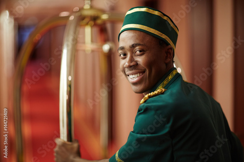 Smiley young bellboy pushing cart with luggage