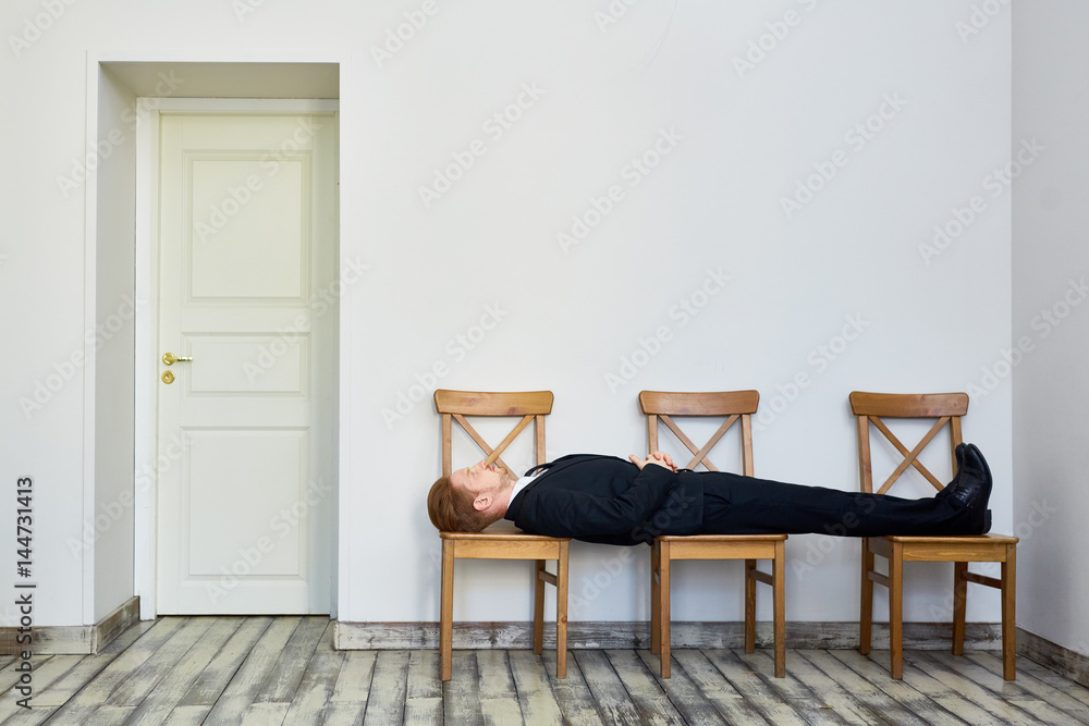 Fototapeta premium Portrait of young red haired business man bored and lying on chairs in waiting room outside office