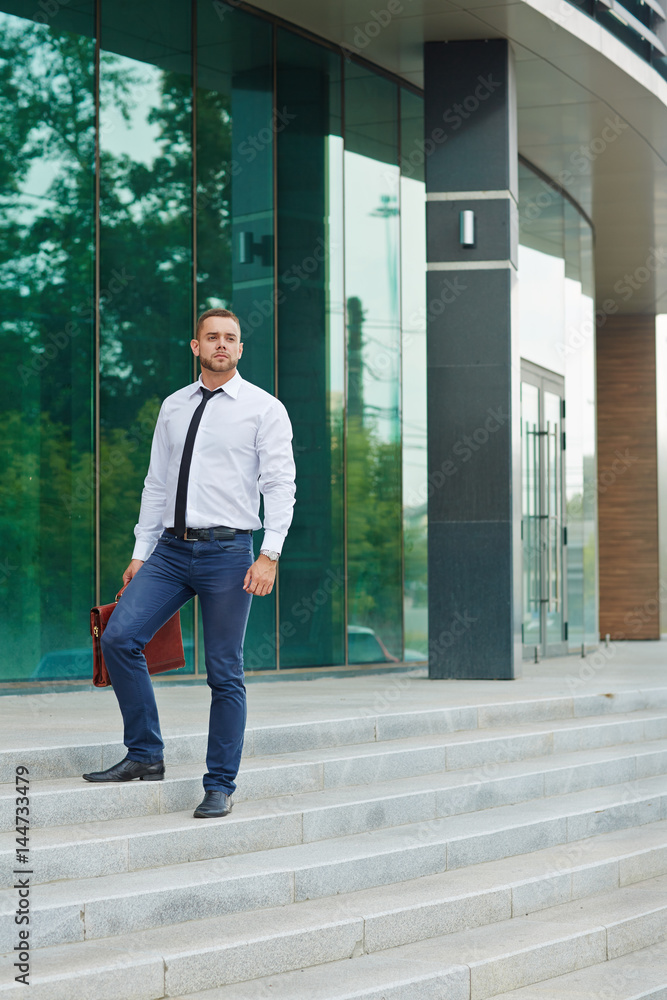 Portrait of handsome young man standing on steps outside modern office building with glass fronts, looking confident