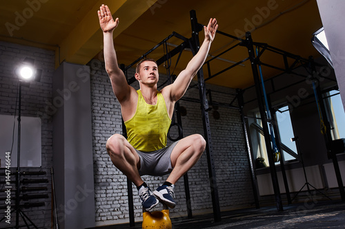Young athletic man doing exercises in cross fit gym while crouching on two legs on the kettlebell. © Aleksey