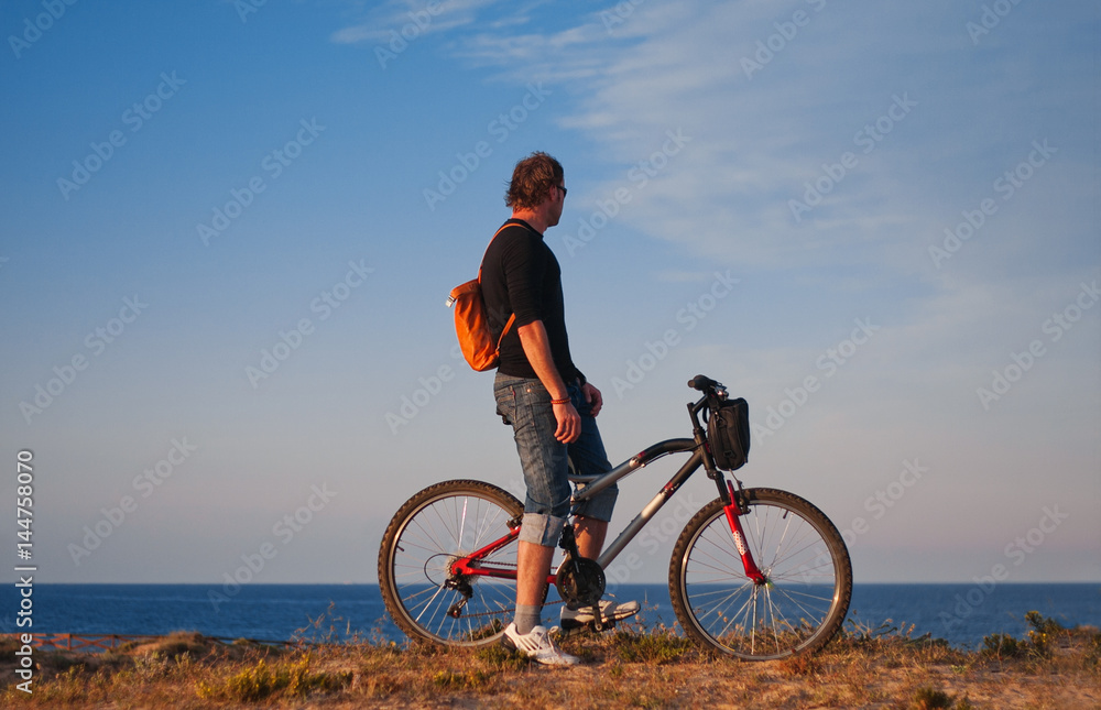 Fototapeta premium A cyclist on a sunset background by the sea. Bicycle tour along the seashore. Evening bike tour. Bright sunset against the background of the Mediterranean Sea 