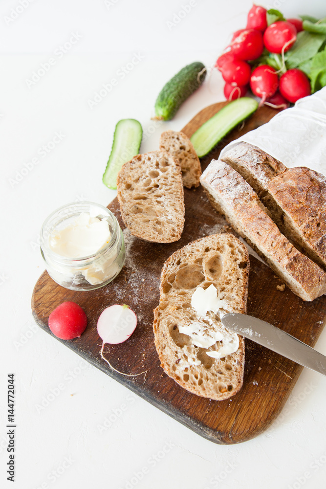 Sliced loaf of homemade bread, with salt, radish, herbs and butter on a white background.