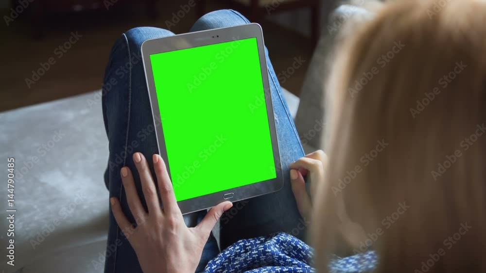 Young woman in blue jeans sitting on couch uses tablet pc with pre