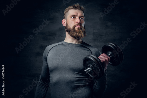 Sporty bearded male in a grey sportswear holds dumbbell.