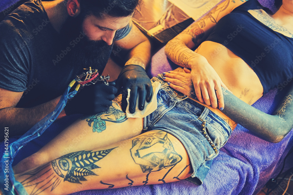 Obraz premium The bearded tattoo artist makes a tattoo on a woman's leg in an ink saloon.