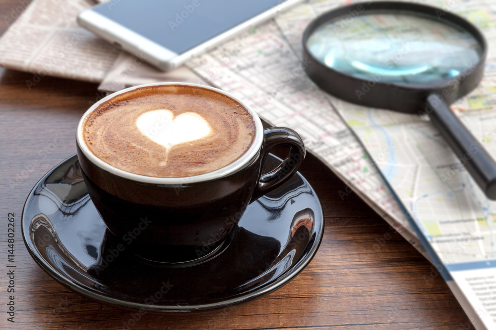 Coffee cup of latte art beside with smartphone, maps and magnifier on wooden table