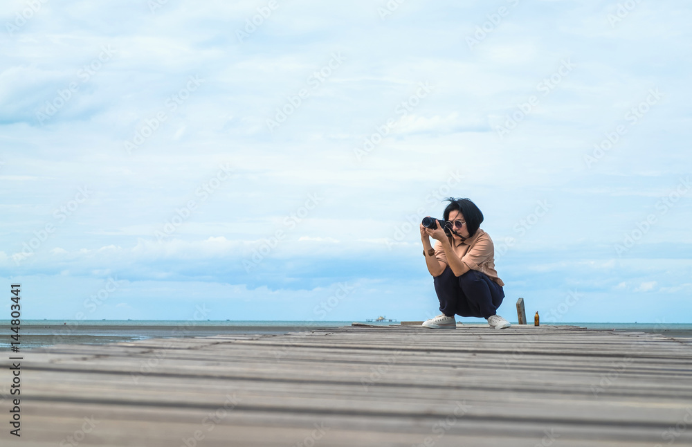 Obraz premium Asian woman traveler take photo of seascape on wood bridge with blue sky and ocean at background,low angel view