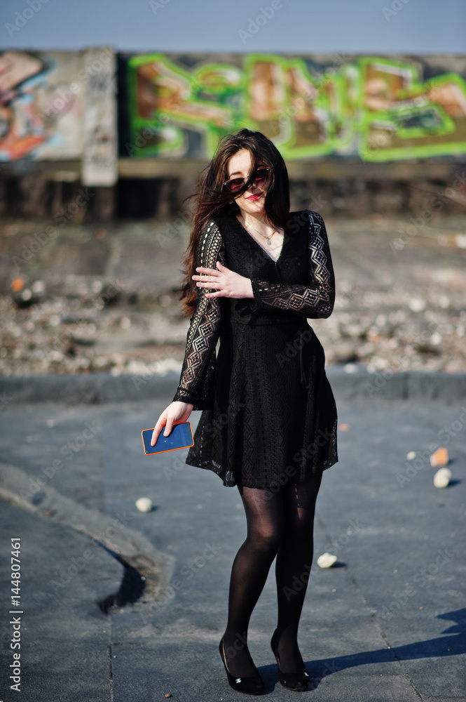 Fototapeta premium Portrait brunette girl with red lips and orange mobile phone at hands, wearing a black dress, sunglasses posed on the roof. Street fashion model.