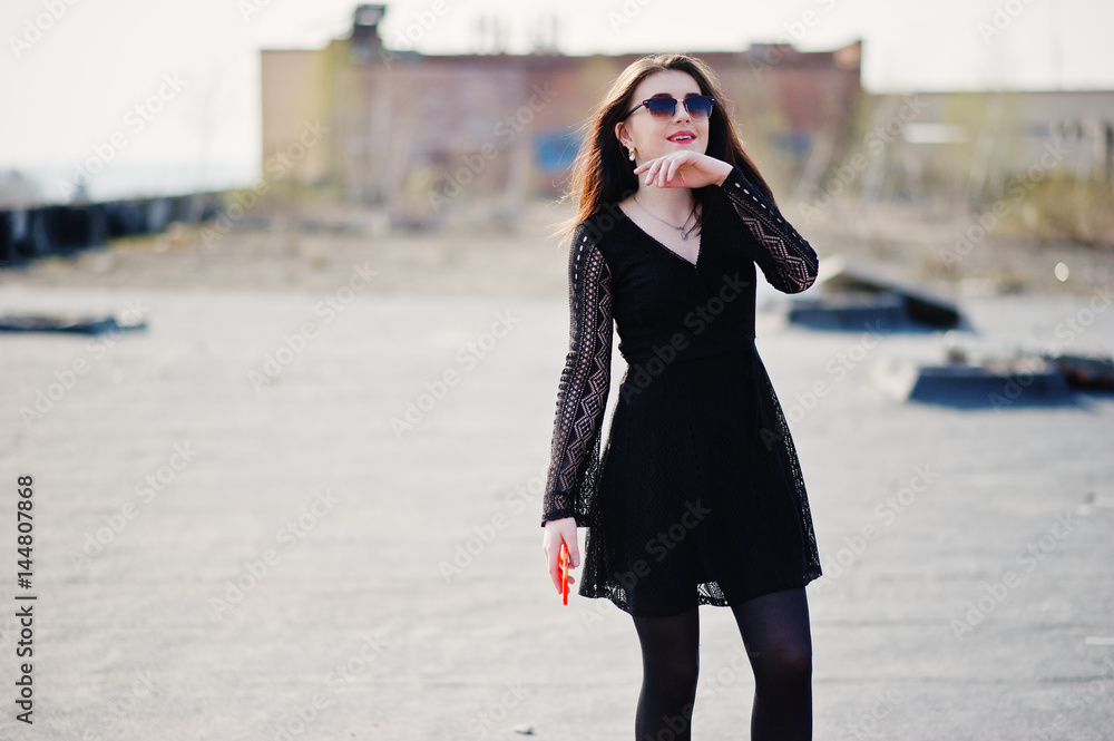 Portrait brunette girl with red lips and orange mobile phone at hands, wearing a black dress, sunglasses posed on the roof. Street fashion model.