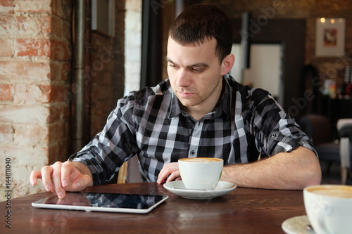 A successful young man in a dark plaid shirt in the cafe does business. Young hipster holding a smartphone, tablet computer. Office worker on lunch. business meeting. send a message.