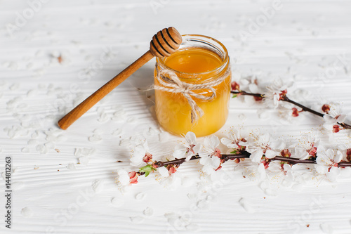 Bank of honey, spoon and blossom branch on a white wooden background.