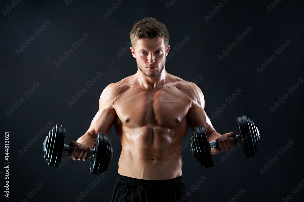 Handsome young fierce muscular male athlete with stunning sexy body and perfect abs looking confidently to the camera lifting heavy weights on black background motivation determination effort.