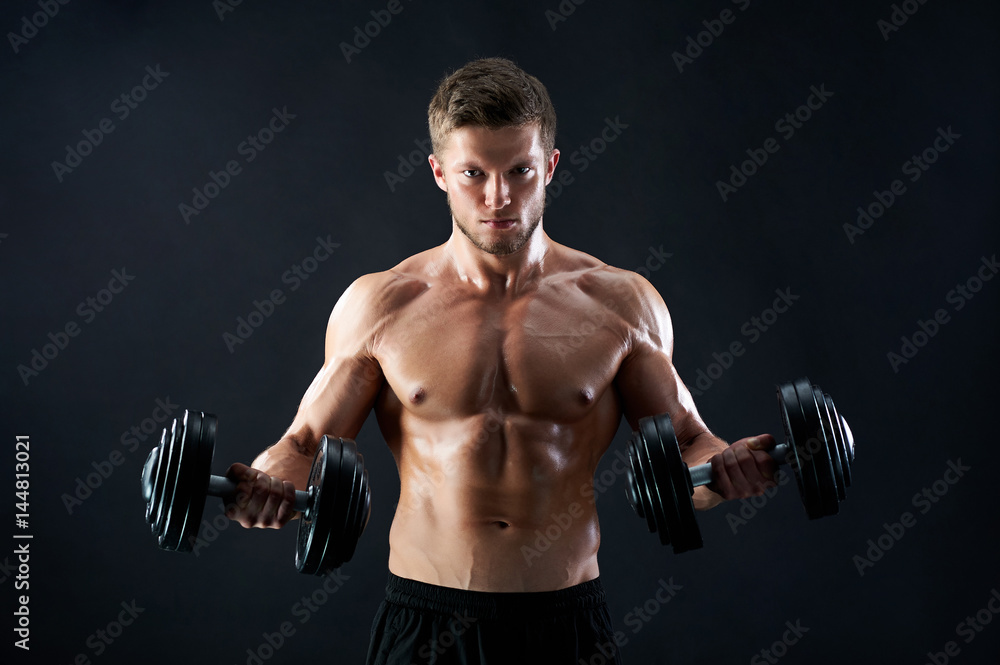 Handsome young fierce muscular male athlete with stunning sexy body and perfect abs looking confidently to the camera lifting heavy weights on black background motivation determination effort. Handsome young fierce muscular male athlete with stunning sexy body and perfect abs looking confidently to the camera lifting heavy weights on black background motivation determination effort.