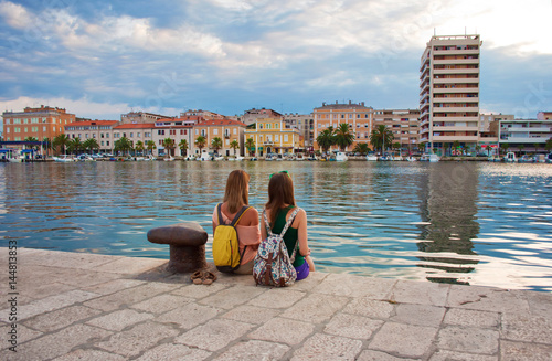 Two young girls with backpacks sitting on the stone pavement near the sea shore looking at the marine and pretty little colorful houses. Town of Zadar, Croatia