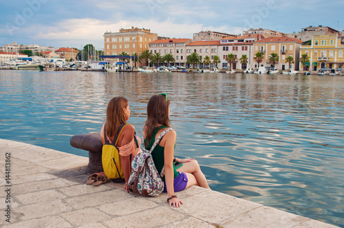 Two young girls with backpacks sitting on the stone pavement near the sea shore looking at the marine and pretty little colorful houses. Town of Zadar, Croatia