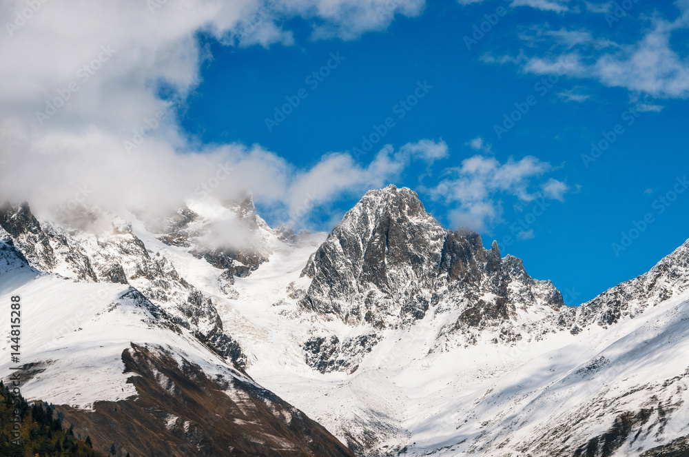 Fototapeta premium Snow covered mountains and their peaks in the clouds against the blue sky in Georgia. Greater Caucasus Mountains