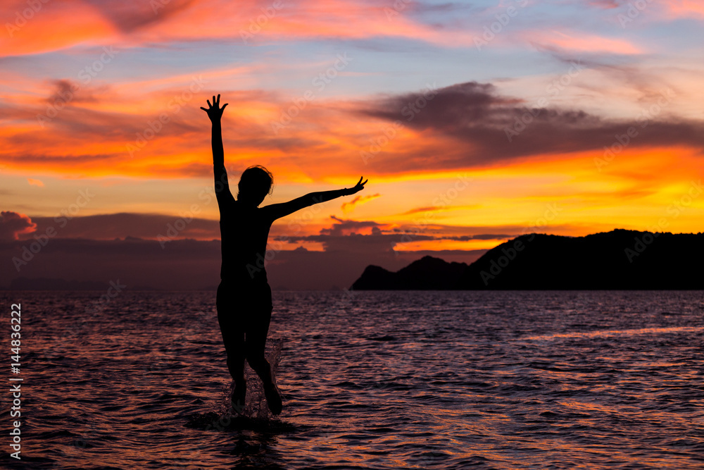 young woman silhouette happily jumping on beach on sunset background