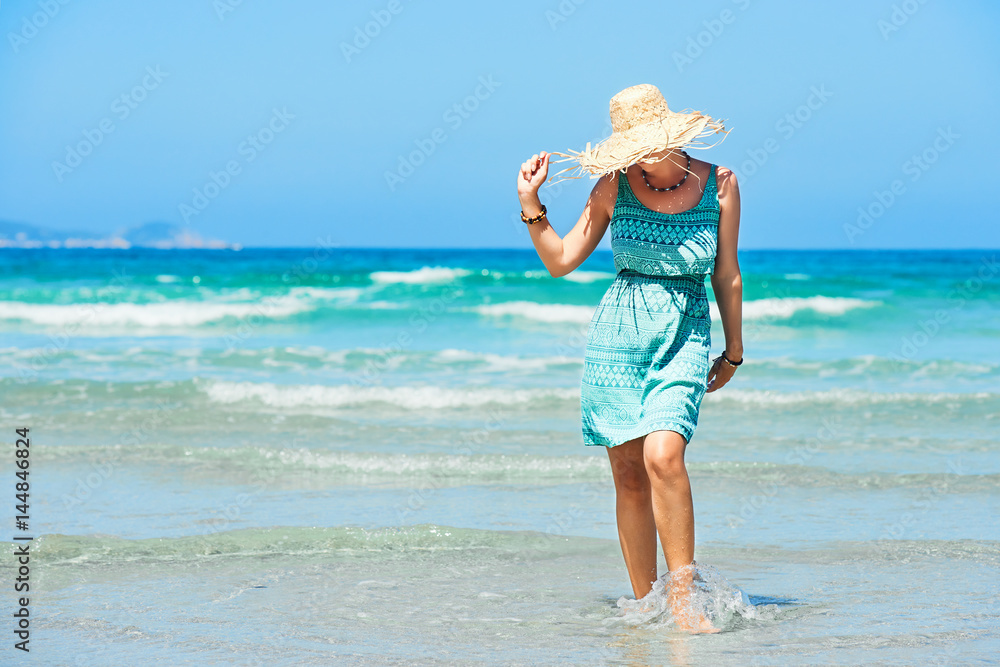 Young woman in summer dress and straw hat walking on beach.