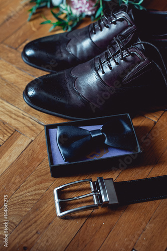 Close up of modern man accessories. Black bowtie, leather shoes, belt and wedding bouquet on wood rustic background. Set for formal style of wearing