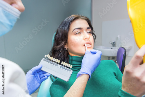Fototapeta Naklejka Na Ścianę i Meble -  Stomatologist holding tooth color chart. Young woman looking into mirror. Teeth whitening at dental clinic.