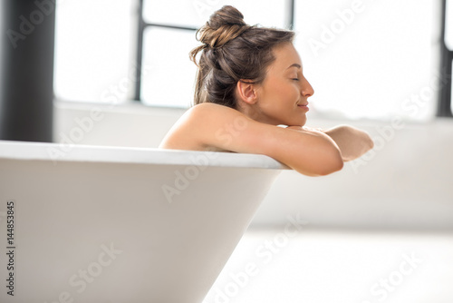 Beautiful young woman relaxing lying in the bathtube at the bright bathroom with windows