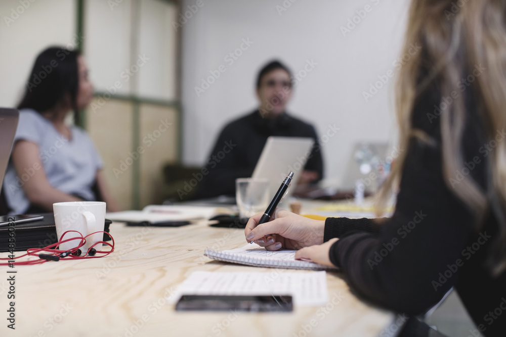 Midsection of young woman writing at desk while colleagues sitting in background Midsection of young woman writing at desk while colleagues sitting in background