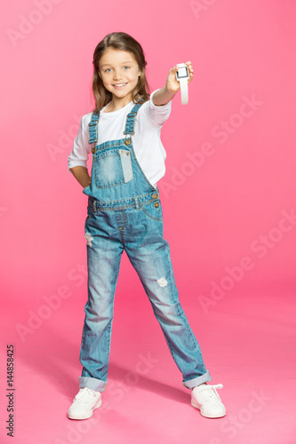Adorable little girl in denim overalls holding smartwatch and smiling