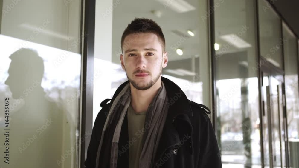 Handsome Caucasian young male in street outerwear standing near shop-window and smiling at camera in slowmotion Handsome Caucasian young male in street outerwear standing near shop-window and smiling at camera in slowmotion