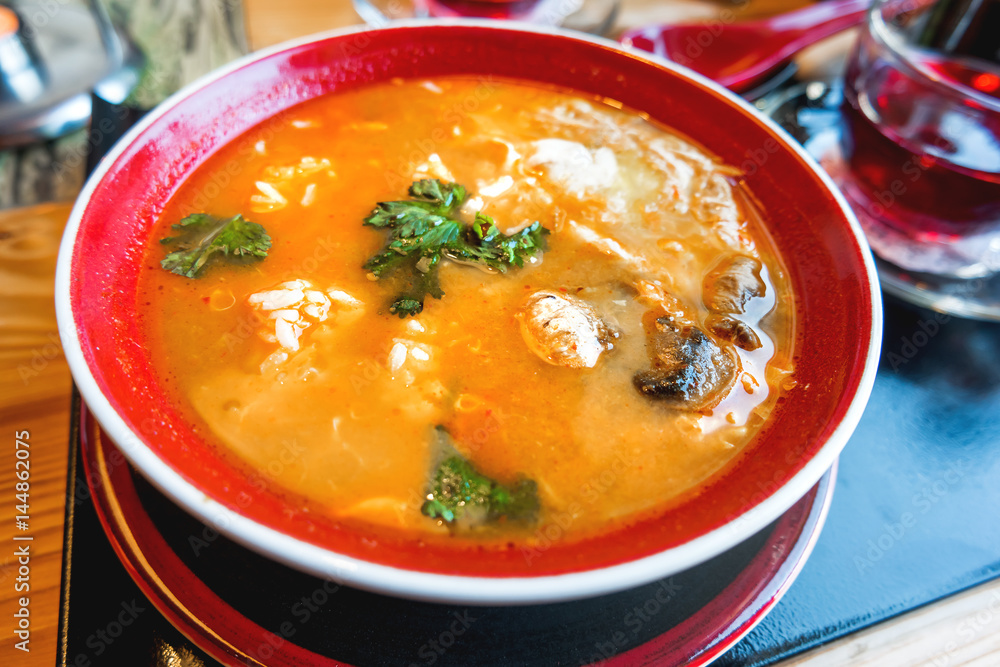 Asian cuisine - Tom kha kai or Thai coconut soup with chicken meat, rice, mushrooms, ginger and lemongrass in red bowl. Close up. Asian cuisine - Tom kha kai or Thai coconut soup with chicken meat, rice, mushrooms, ginger and lemongrass in red bowl. Close up.