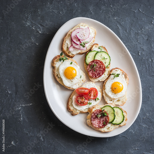 Variety of mini sandwiches with cream cheese, vegetables, quail eggs and salami. Sandwiches with cheese, cucumber, radish, tomatoes, salami, quail eggs on a dark background, top view