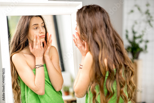Young woman looking at the mirror troubled with wringles on the face standing in towel at the bathroom