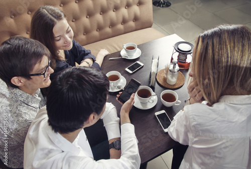 Group of young people from two couples of guys and girls have a tea time in the cafe and looking on the smartphone absorbedly