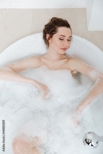 Relaxed pretty young woman lying in bathtub with foam