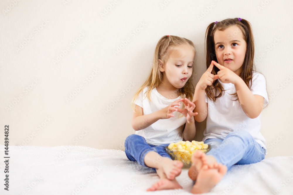 Portrait of beautiful young sisters eating popcorn at home. Two children eat popcorn against the white background Portrait of beautiful young sisters eating popcorn at home. Two children eat popcorn against the white background