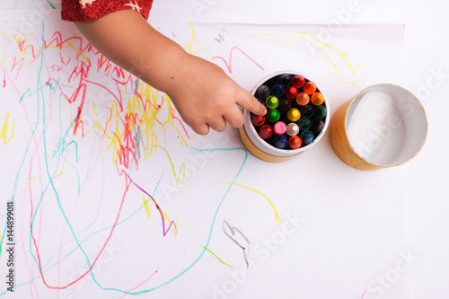 A hand of baby selecting a color of crayon. Colorful crayon are in a round box. With line and shape on the white paper