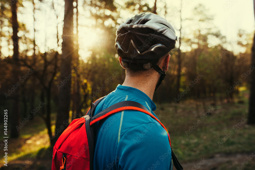 Close up shot from back of handsome young bearded man cyclist wearing protective helmet, blue t-shirt and red backpack, looking away, thinking about route and cycling. Travel concept. Sport lifestyle. Close up shot from back of handsome young bearded man cyclist wearing protective helmet, blue t-shirt and red backpack, looking away, thinking about route and cycling. Travel concept. Sport lifestyle.