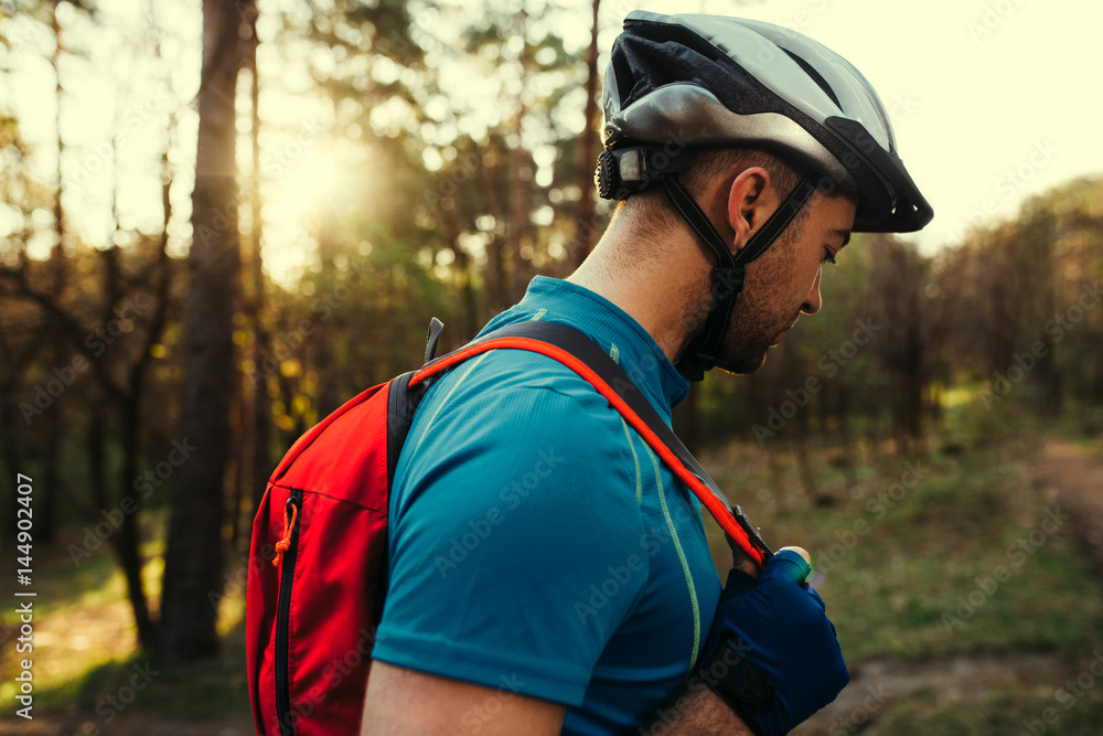 Beautiful close up shot of handsome young bearded man cyclist wearing protective helmet, blue t-shirt and red backpack, looking down, thinking about route and cycling. Travel concept. Sport lifestyle. Beautiful close up shot of handsome young bearded man cyclist wearing protective helmet, blue t-shirt and red backpack, looking down, thinking about route and cycling. Travel concept. Sport lifestyle.