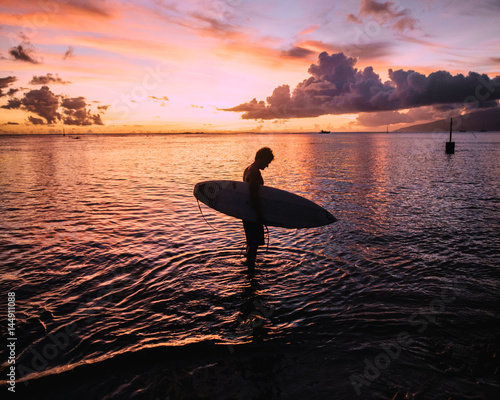 Man by shoreline holding surfboard at sunset, Tahiti, South Pacific  photo