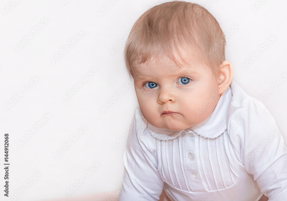 Beautiful girl with blue eyes in white dress stands on all fours on the floor and seriously looking at the camera. Portrait of a baby in 9 months